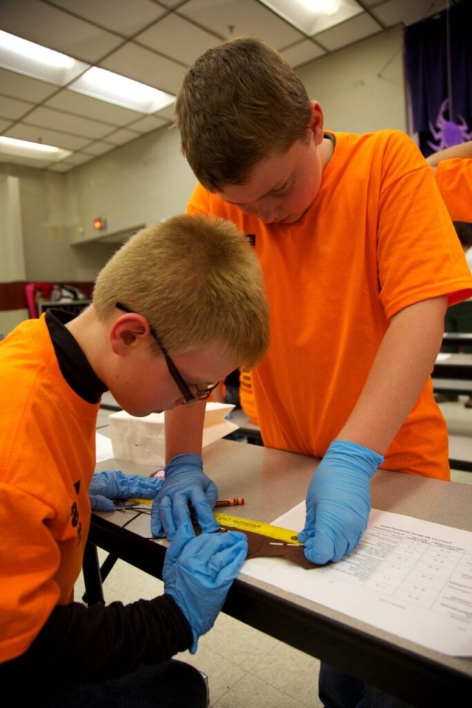 Boys creating a visual project with crayons and a ruler in a school cafeteria